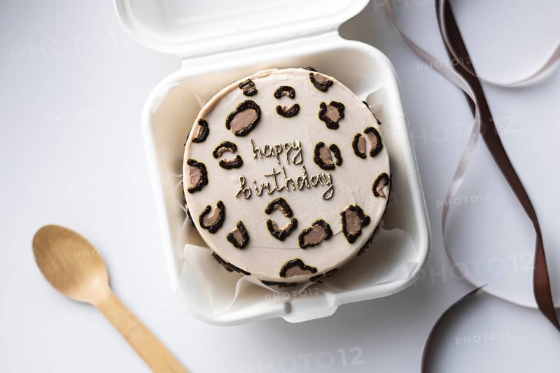 Top view of a mini bento birthday cake styled with a leopard print design and a Happy Birthday message, in a white takeout box, accompanied by a wooden spoon and satin ribbon