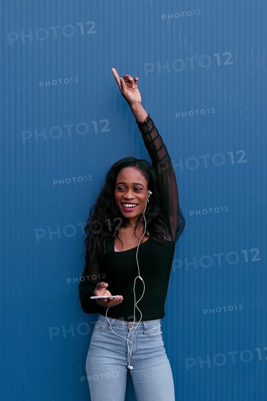 A young woman in casual attire, listening to music with earphones, raises her hand in excitement. She stands against a vibrant blue background, carrying a smartphone
