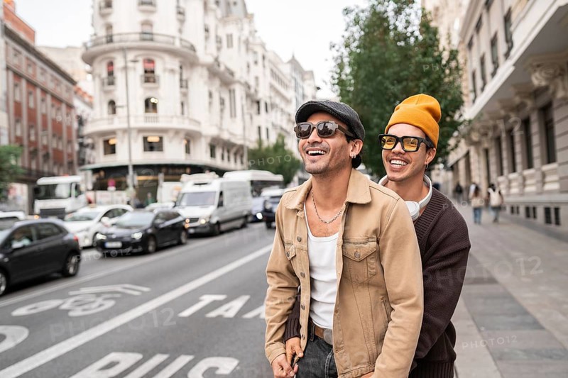 A joyful colombian gay couple enjoying a casual city walk. Both are wearing sunglasses and showcasing happiness and connection against an urban backdrop of architecture and vehicles