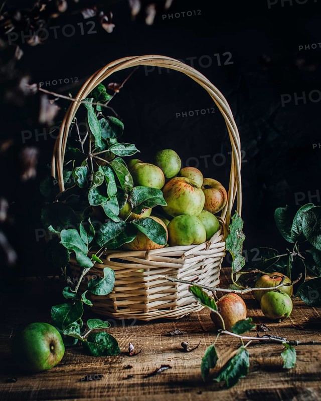 A rustic wicker basket filled with fresh green apples sits on a wooden table. Surrounded by leaves and twigs, the scene evokes a cozy, autumn harvest atmosphere