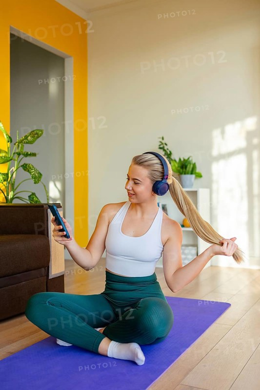 A woman in active wear sits on a yoga mat at home, wearing headphones and looking at her phone. She is relaxed, with sunlight and plants creating a peaceful ambiance