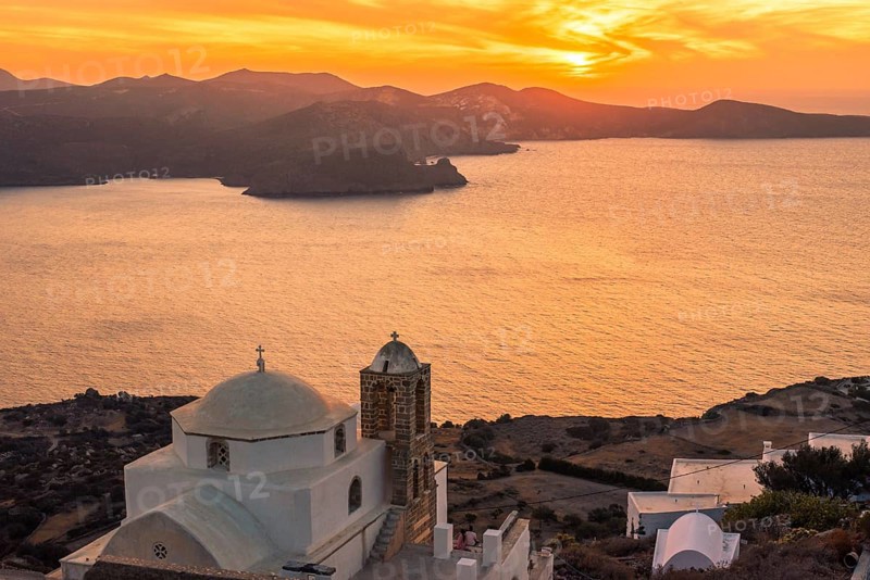 A serene sunset over the Greek coastline, featuring a classic white church with a dome in Plaka, Milos island. The sun casts a golden hue over the sea and surrounding rugged mountains