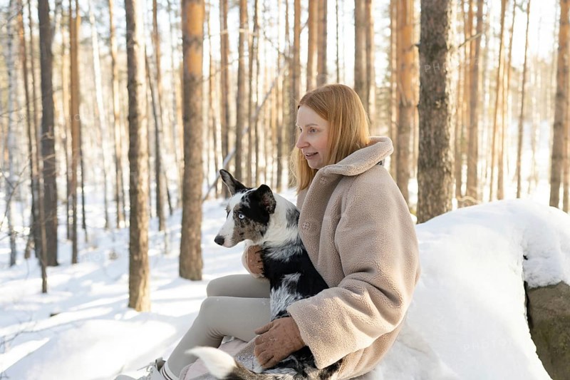 A redhead woman walks through a snowy forest with her Border Collie. The serene winter scenery complements the calm interaction between her and the dog, capturing a moment of tranquility and companionship in nature