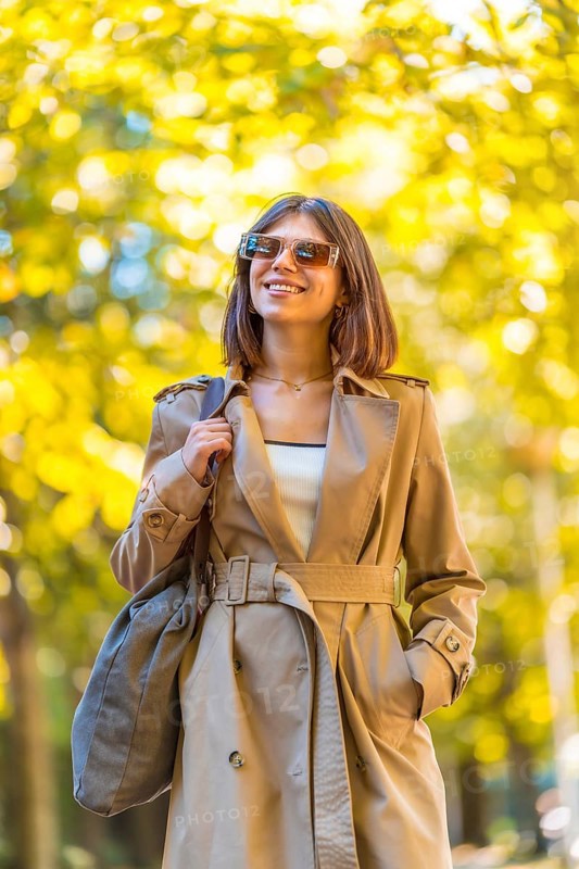 A woman wearing a tan trench coat and sunglasses is walking down a path. She is smiling and holding a handbag. Autumn or winter concept in the city