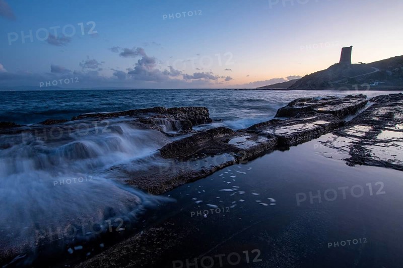 The serene sunset over the rocky coast of Tarifa, Cadiz, casts a soft glow on the water. Calm waves splash against the rocks, reflecting the peaceful ambiance of the scene