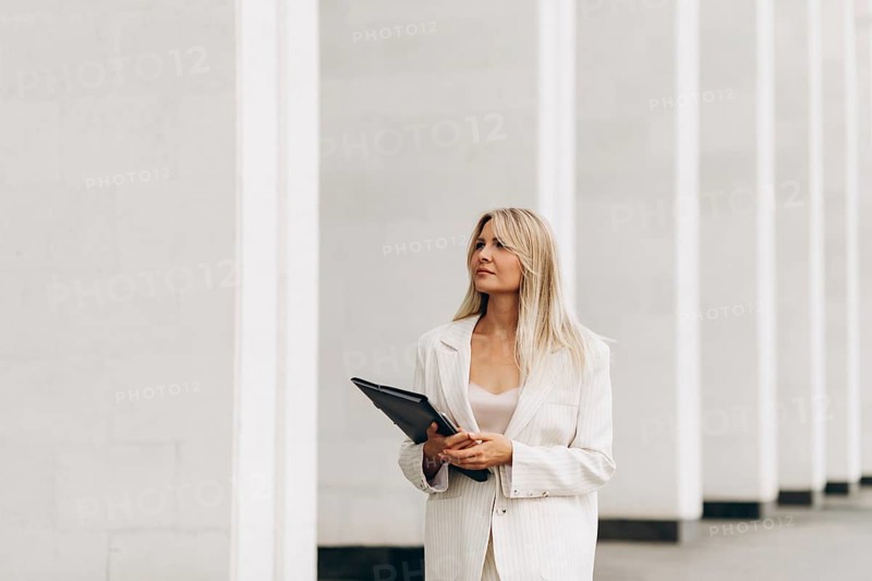 A professional woman holding a folder walks confidently beside a modern architectural structure. She is dressed in a stylish suit, exuding confidence and focus