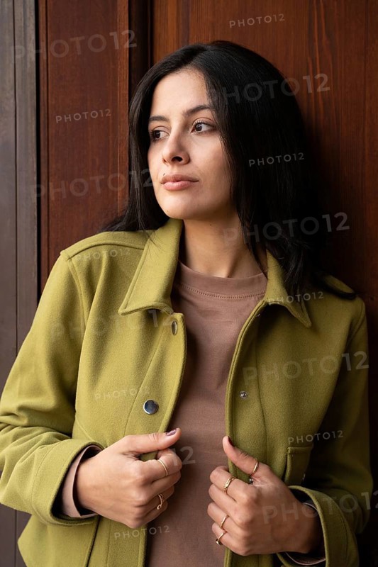 A woman stands against a wooden background, wearing a green jacket and brown top. Her gaze is thoughtful and contemplative, creating a mood of introspection