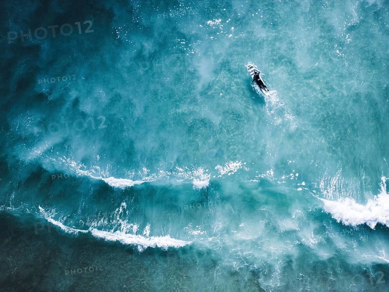 Aerial shot capturing a surfer navigating the clear blue waves of Portugal. The image emphasizes the natural beauty and dynamic motion of the ocean landscape