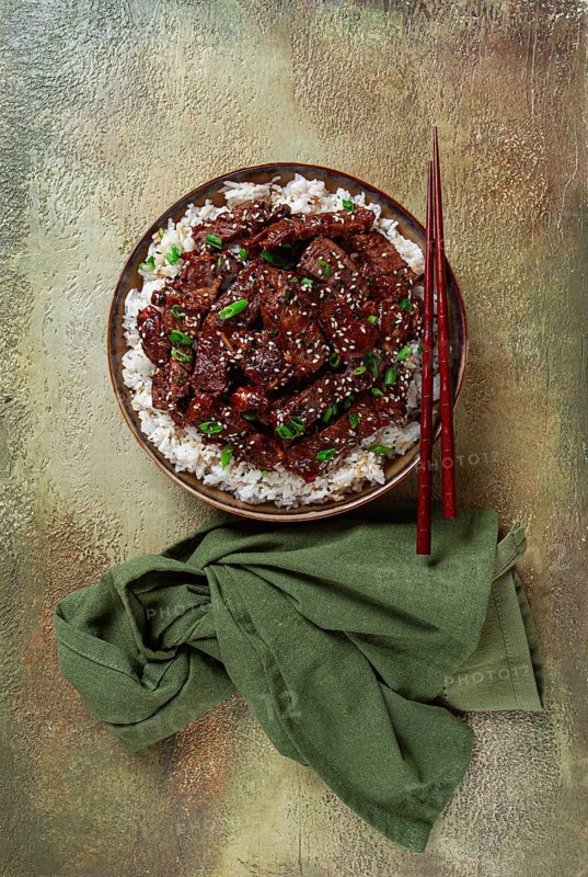 Crispy beef, with green onions and sesame seeds, on a rice pad, in a bowl with chopsticks, on a textured surface, no people