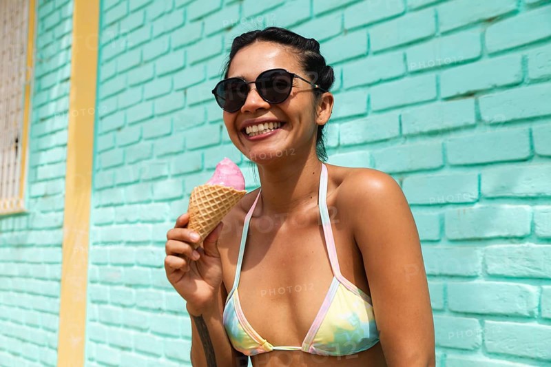 A joyful young Latin woman savors an ice cream cone under the sun in Mancora, Peru. Her bright bikini and sunglasses add to the relaxed beach town vibe