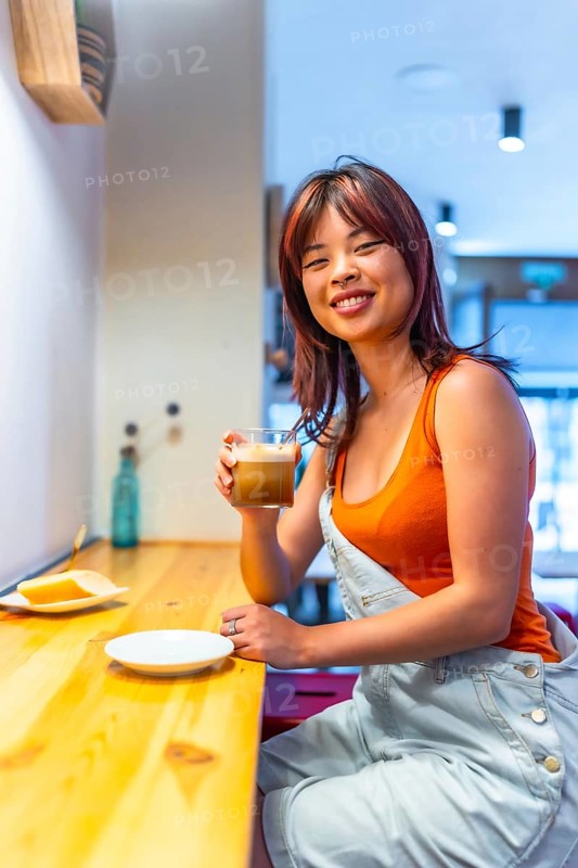 Vertical photo of a young casual chinese smiling woman enjoying morning coffee in a cafeteria