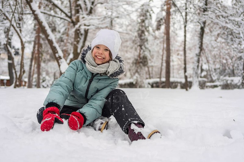 A cheerful child dressed warmly in a snowy forest, enjoying a winter day. The child wears a winter jacket, hat, and gloves, surrounded by snow-covered trees