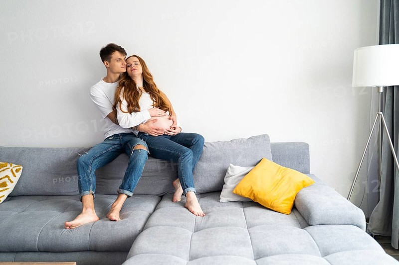 A young couple expecting a baby sits lovingly on a comfortable sofa, embracing each other with warmth and joy in a bright, minimalist living room setting