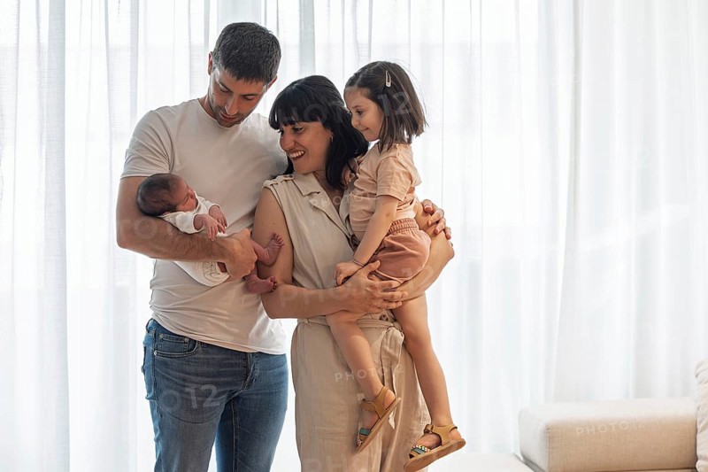 A joyful young family is gathered indoors, celebrating the arrival of their newborn baby. The big sister stands happily, sharing in the love and warmth of this special moment