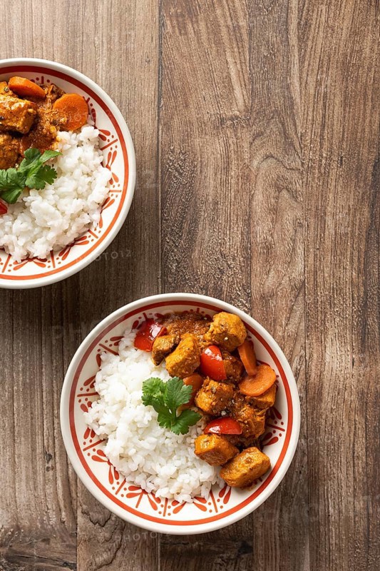 From above, two bowls of Hungarian pork goulash served with rice, garnished with a parsley leaf on a wooden surface. The dish features chunks of pork, carrots, and tomatoes in a savory sauce