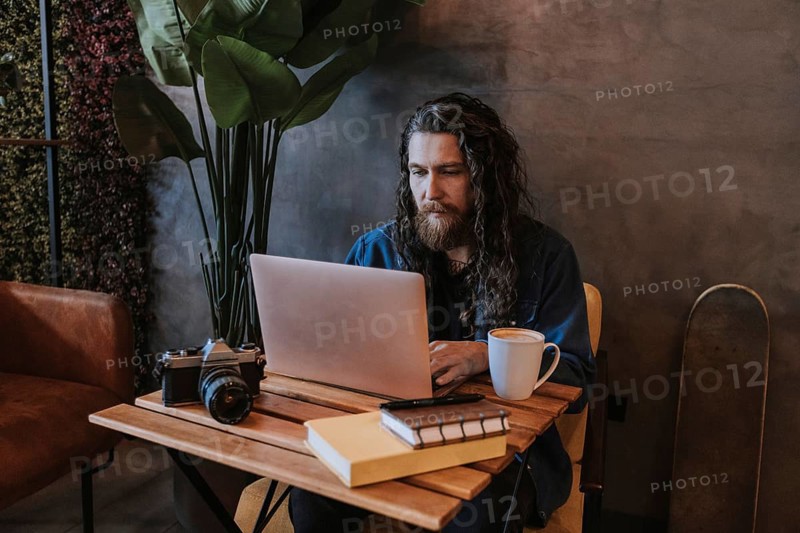 A man with long hair and a beard types on a laptop in a cozy cafe. A vintage camera, books, and a mug of coffee are on the wooden table beside him, adding to the artistic atmosphere