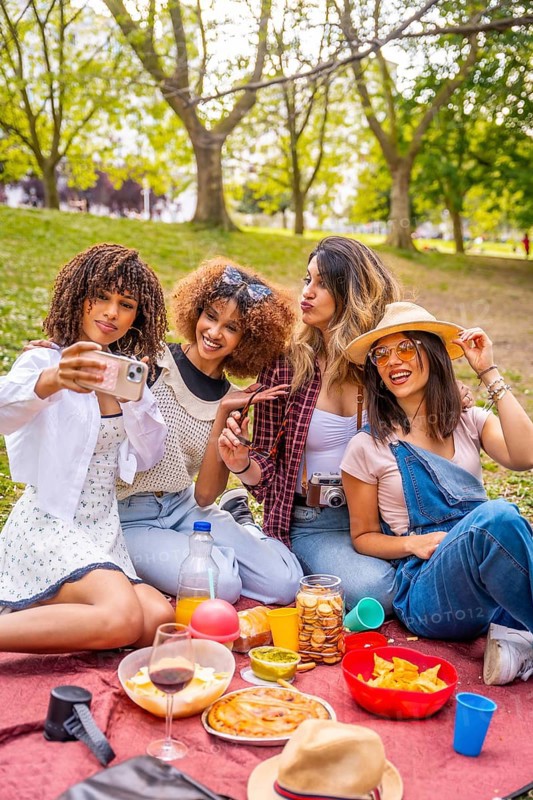 Four diverse female friends are enjoying a picnic in a park, taking a selfie with a smartphone