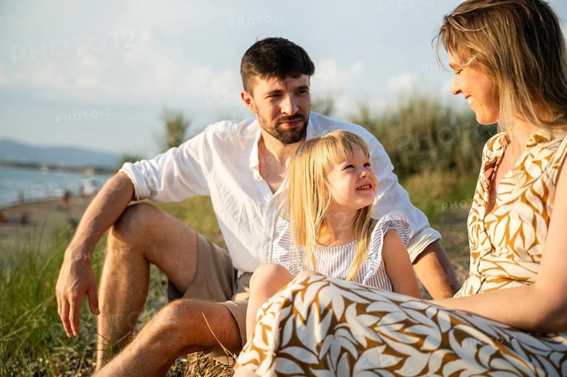 A loving family sits together on a peaceful beach in Italy, enjoying the warm summer air. The parents and their daughter share smiles as they take in the beautiful coastal scenery