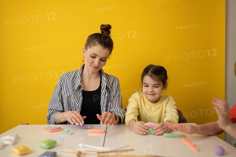 A woman and child create colorful clay shapes at a white table against a vibrant yellow background, encouraging creativity and learning through play in a warm environment