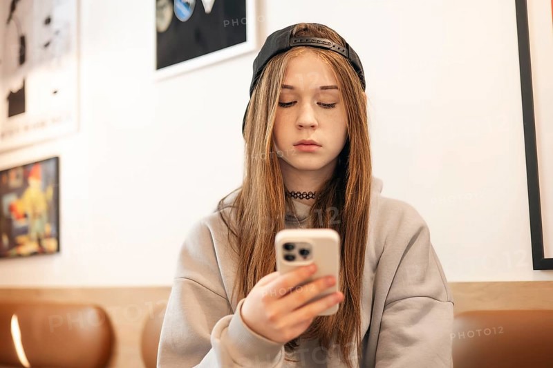 A teenage girl in a casual hoodie and cap sits indoors, focused on her smartphone. The background features modern art, emphasizing a contemporary youthful setting