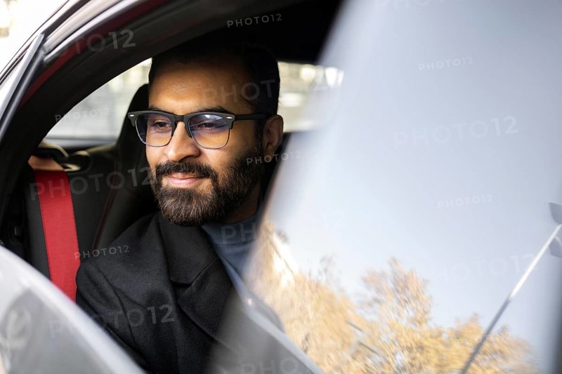 An Indian man dressed in a dark winter coat and glasses is seen smiling gently while driving his car, conveying a sense of contentment and comfort