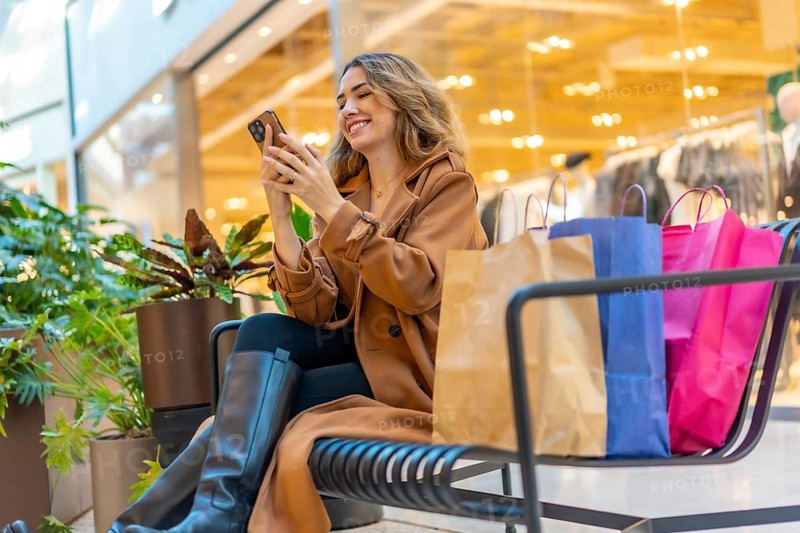 Happy woman taking a break from shopping at the mall, sitting on a bench with shopping bags and using her mobile phone, actively engaging with online content