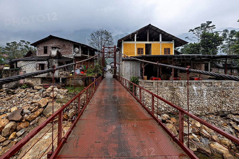 A metal bridge connects two older buildings, one of them yellow, under a grey cloudy sky, A bridge over a river in the countryside near Sa Pa in Vietnam