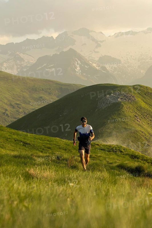 A lone runner navigates a picturesque trail through vibrant green mountains, wearing a hat and sunglasses under a cloudy sky, embodying the spirit of outdoor adventure