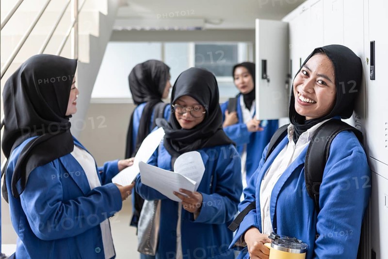 Asian Muslim students in hijabs and uniforms gather in a bright school hallway, engaging with each other and holding educational materials, showcasing the back to school spirit