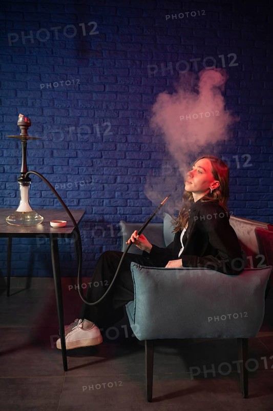 A woman enjoys a hookah in a modern lounge against a blue brick wall. Smoky clouds create a serene and relaxing atmosphere. Stylish and comfortable setting for unwinding