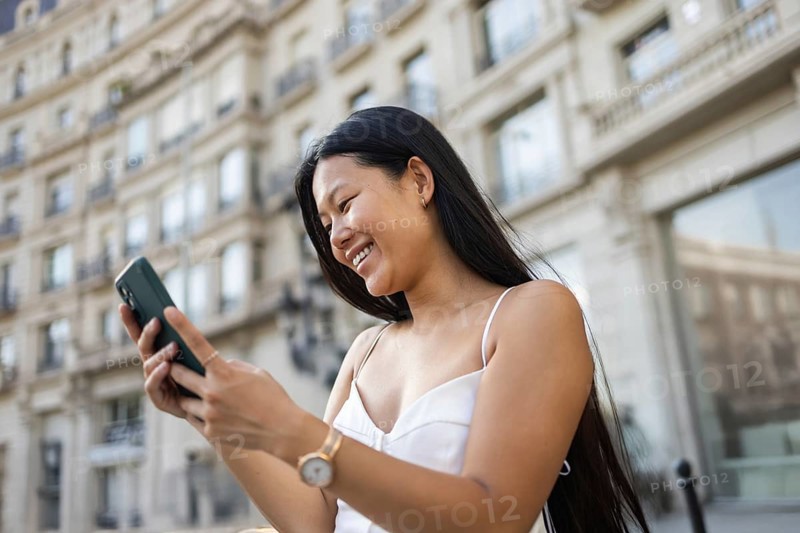 A young Chinese woman smiling while using her smartphone on a sunny summer day in the city. The urban backdrop enhances her lively and joyful expression