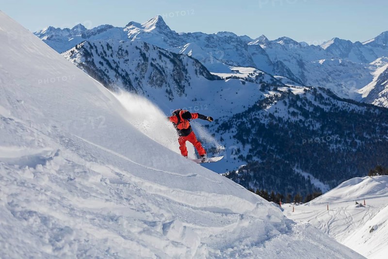 A snowboarder wearing bright red gear carves down a snowy mountain slope. The backdrop features stunning snow covered peaks under a clear blue sky