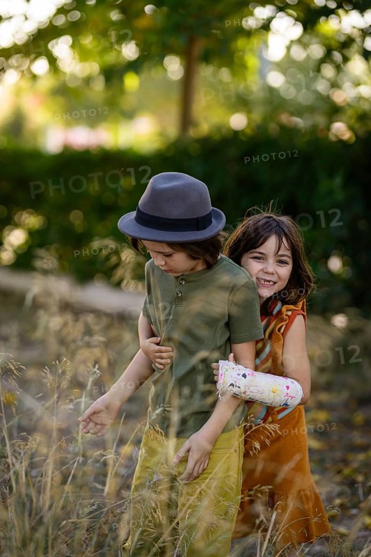 Two children playfully enjoy a sunny day outdoors. One wears a hat, while the other has a colorful cast. The background is filled with lush greenery and sunlight
