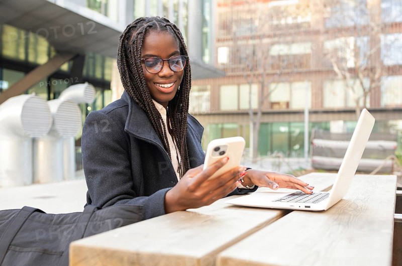 A young African American businesswoman with braids is smiling as she uses her smartphone and laptop while working remotely outdoors, capturing a moment of work and relaxation