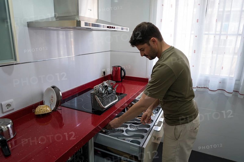 A man is organizing utensils in a modern kitchen. He carefully arranges cutlery in a drawer, demonstrating efficient household management and attention to detail
