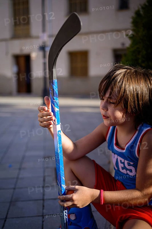 A young child sits on a paved street, holding a vibrant blue hockey stick. The scene captures a moment of quiet focus and passion for the sport in an urban environment