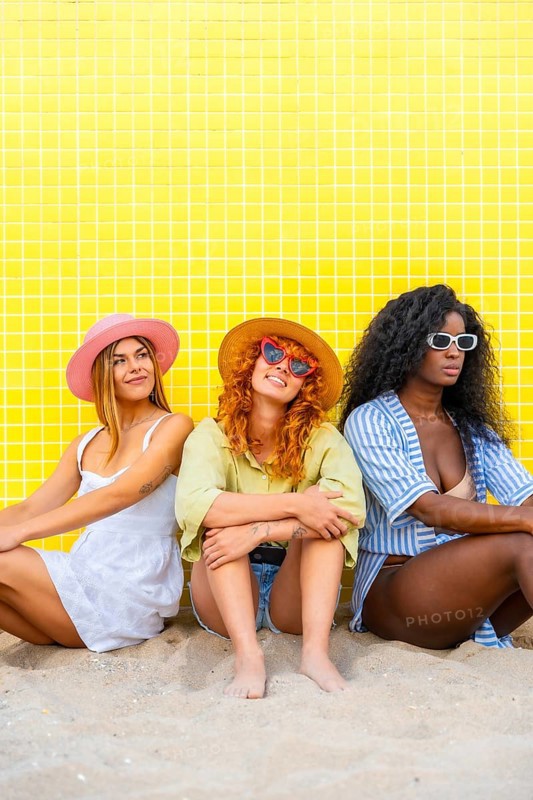 Three young women wearing summer clothes and sunglasses are sitting on the sand against a yellow tiled wall