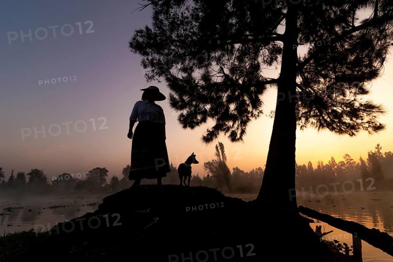 A serene silhouette of a woman and her Xoloitzcuintle dog at sunrise, against the backdrop of the tranquil waters and lush surroundings of Xochimilco, Mexico