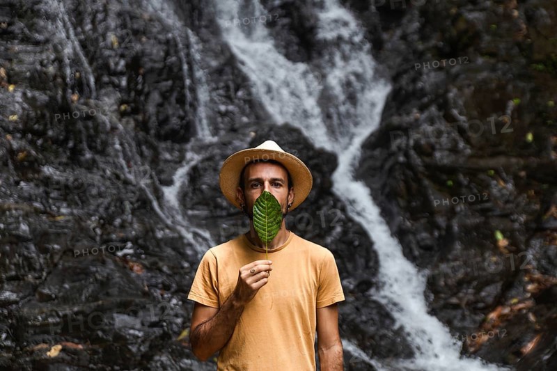 Man from Spain holding a leaf in front of his face, with a waterfall in the Choco Andino rainforest, Ecuador. He is engaged in exploring the rich biodiversity and experiencing ecotourism