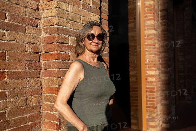 A joyful mature woman in casual attire stands against a brick wall, basking in the warm sunlight. Her smile and sunglasses reflect a relaxed, confident lifestyle