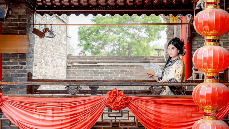 A woman in traditional Qing Dynasty attire, holding a fan, stands gracefully on a decorated balcony in Pingyao, China. The vibrant red lanterns frame the historical scene