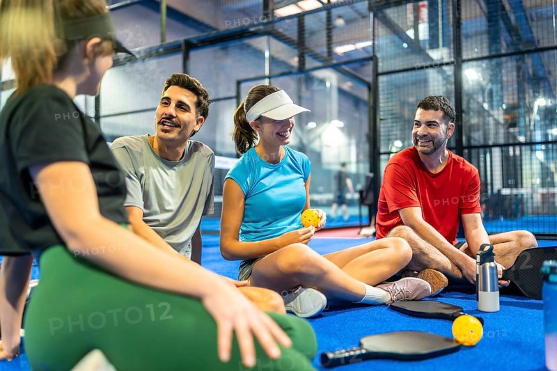 Group of diverse friends taking a break on an indoor pickleball court, smiling and having a social conversation after an active game, enjoying camaraderie and sportsmanship