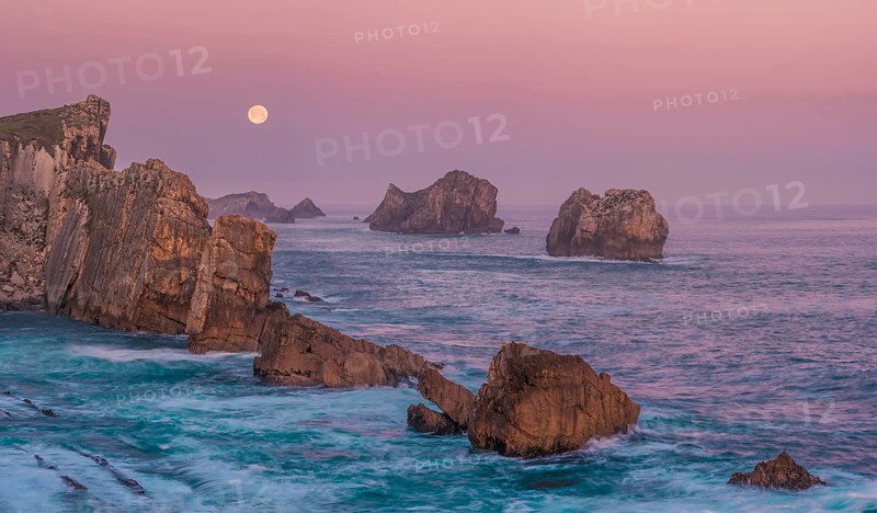 A serene view of rugged sea stacks and cliffs bathed in soft dawn light at Arnia Beach, Costa Quebrada, with the full moon glowing over the tranquil Atlantic horizon