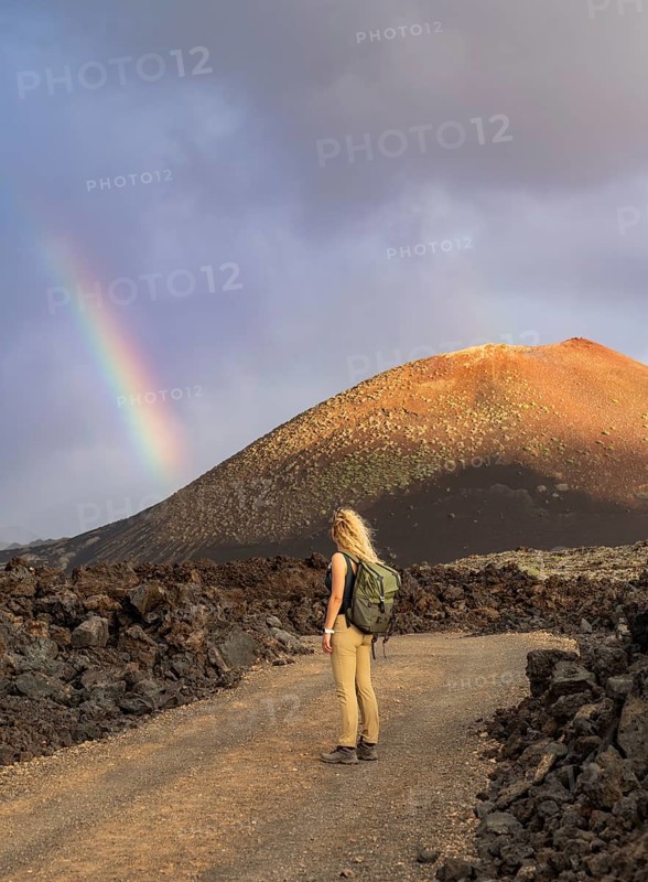 A woman with a backpack stands on a rocky path in Timanfaya National Park, Lanzarote. A vibrant rainbow arcs over the volcanic landscape under a dramatic sky