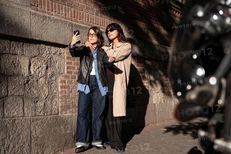 A multiethnic lesbian couple poses for a joyful outdoor selfie, exuding pride and love. The duo stands close, capturing a moment of happiness and unity