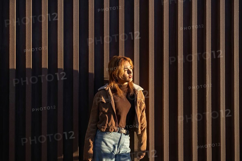 A Mixed-race woman stands against a vertical wooden wall, basking in warm sunlight She wears a casual brown jacket and jeans, embodying a relaxed, pensive mood