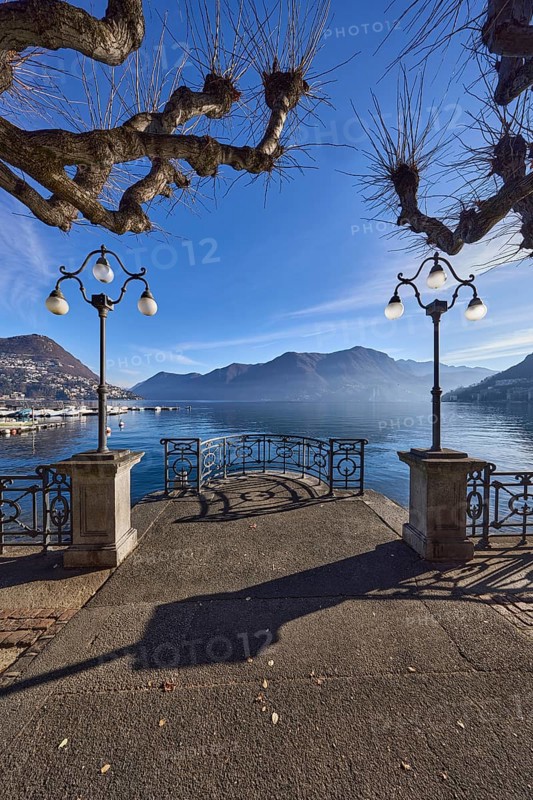 Lake, lakeside promenade, lanterns, trees, railings, mountains, haze, shadow, backlight, Lake Lugano, Lugano, Lugano district, Canton Ticino, Switzerland