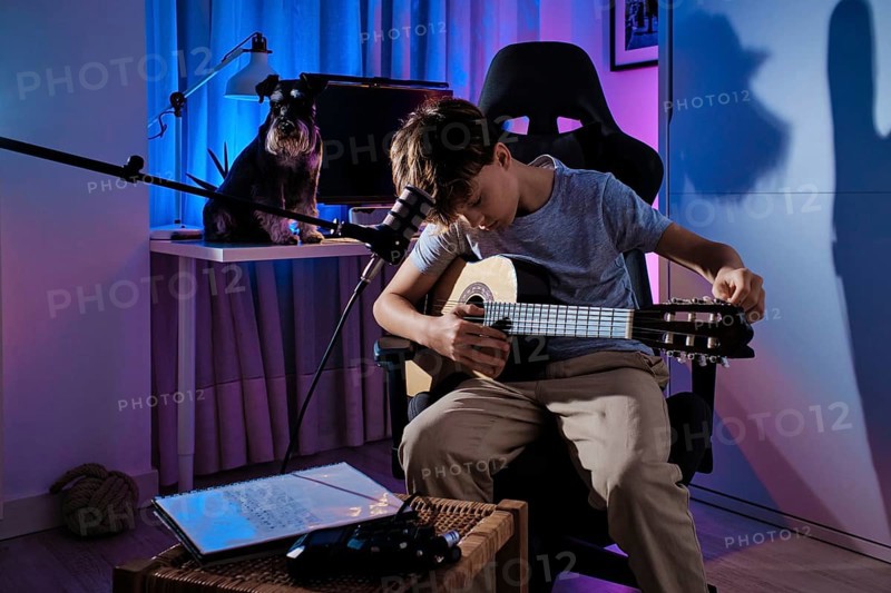 A young boy plays an acoustic guitar in a modern room with colorful lighting. A dog sits nearby on a desk, creating a cozy and creative atmosphere at home