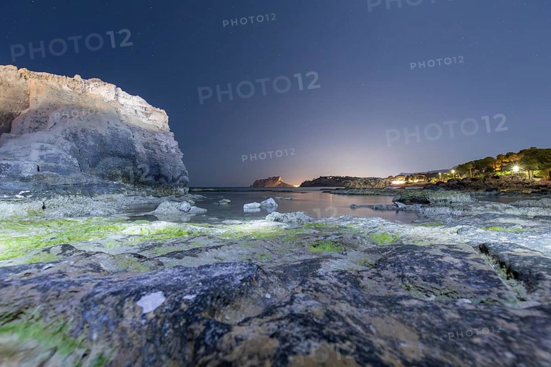 Tranquil Mediterranean shoreline on the Costa Blanca with rocky cliffs and calm sea under a starry night sky. Lights from distant shores reflect on the serene water surface