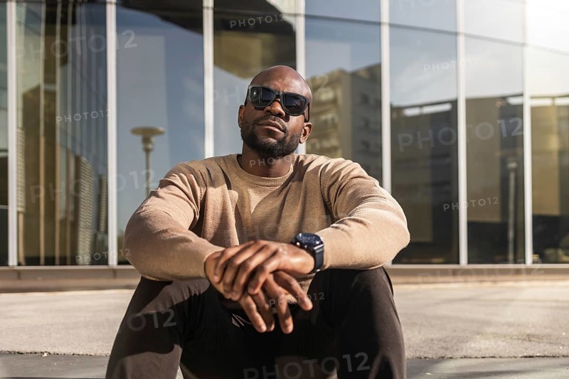 A confident cuban man wearing sunglasses and a casual beige sweater sits outdoors against a modern glass building, enjoying a sunny day with a relaxed and stylish demeanor